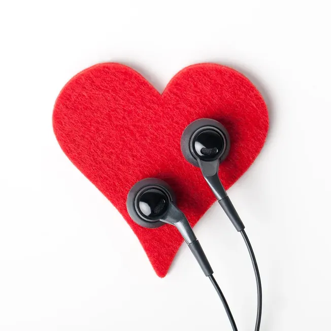 Heart-shaped red plush with black wired earbuds, symbolizing music love and emotional connection, on a clean white background