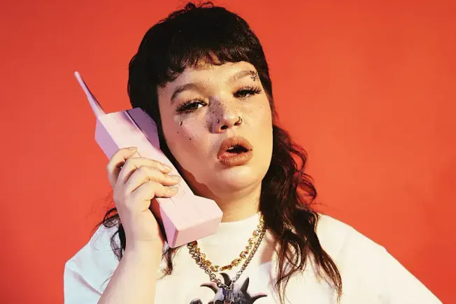 Cute young woman with dark hair and makeup talking on a pink retro phone against a vibrant orange background.