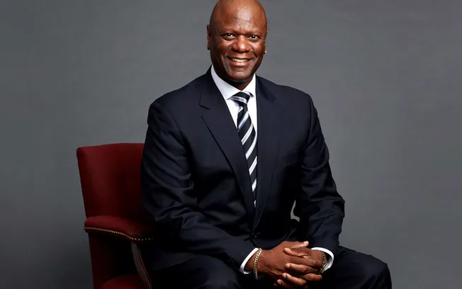 Senior African American man in formal suit sitting on a red chair against a neutral gray background, smiling confidently show