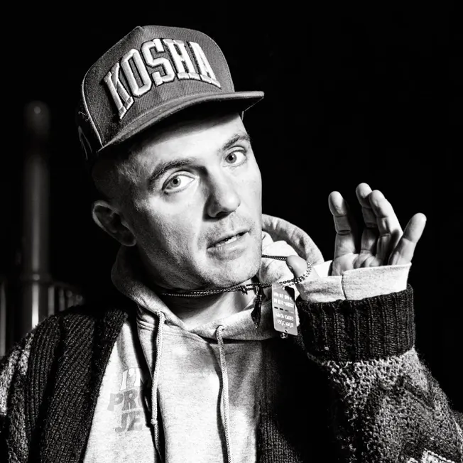 Young man wearing a "KOSH" cap, hoodie, and sweater, making a hand gesture, candid style, black and white photograph, express