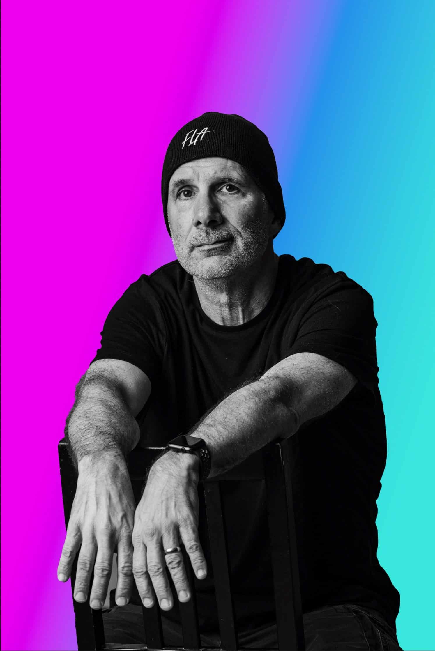 Thought leader in music industry, wearing a beanie and black t-shirt, posed against vibrant pink and blue background for Hypebot feature.