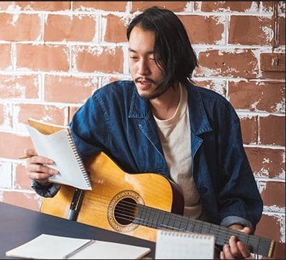 Guitarist practicing in a cozy room with a brick wall background, showcasing music therapy, songwriting, and acoustic guitar skills for musicians.
