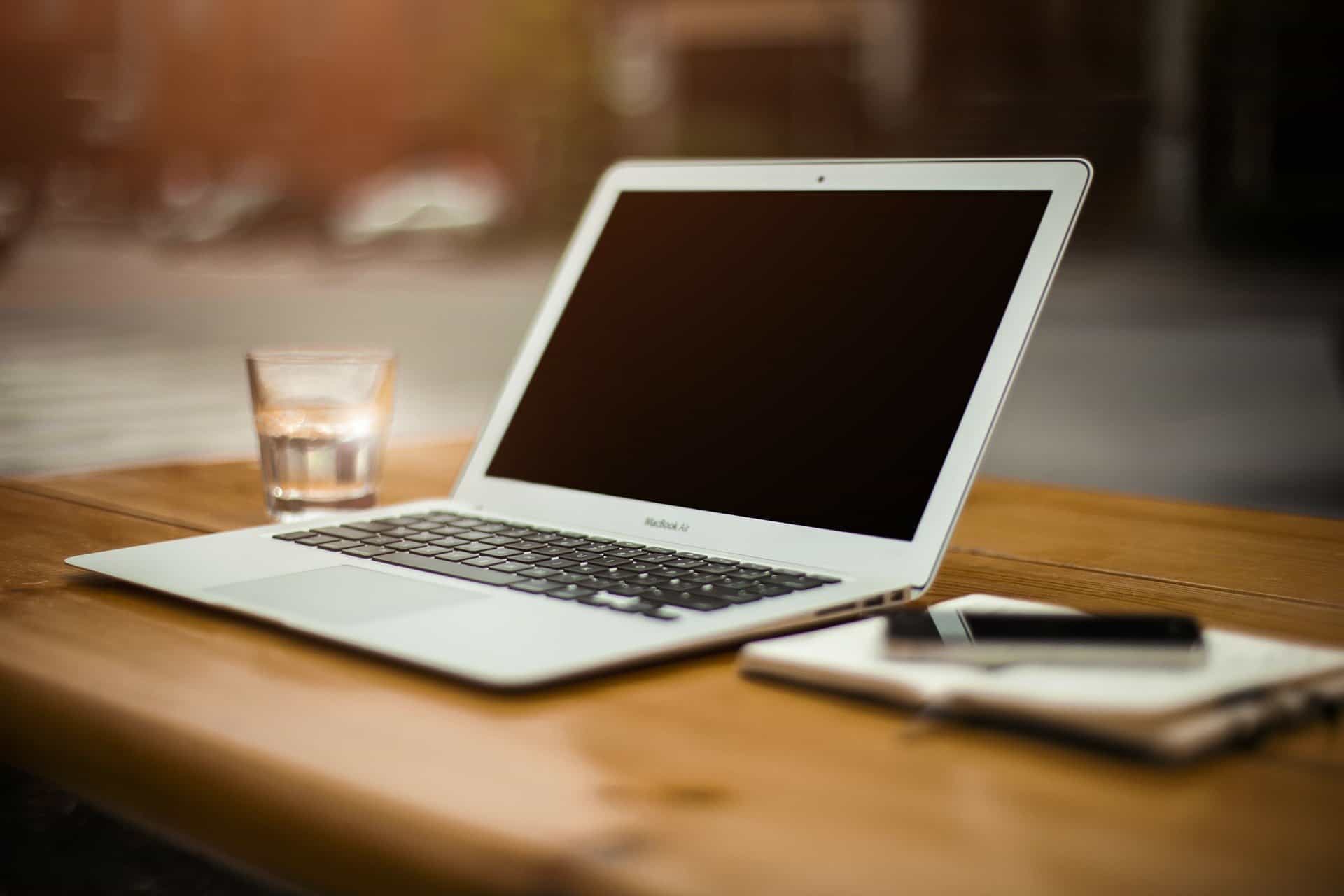 Laptop computer on wooden desk with glass of water, smartphone, and notepad, workspace setup, modern office environment, technology, remote work, digital device, productivity, business workspace.