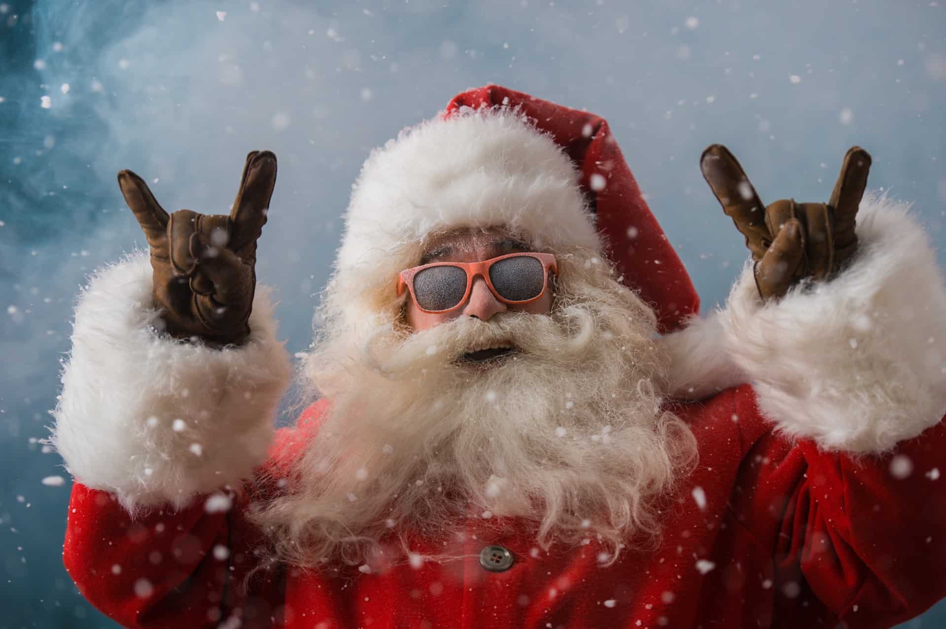 Santa Claus in a winter setting wearing sunglasses, making rock and roll hand gestures amidst falling snow, holiday fun, and festive Christmas spirit.