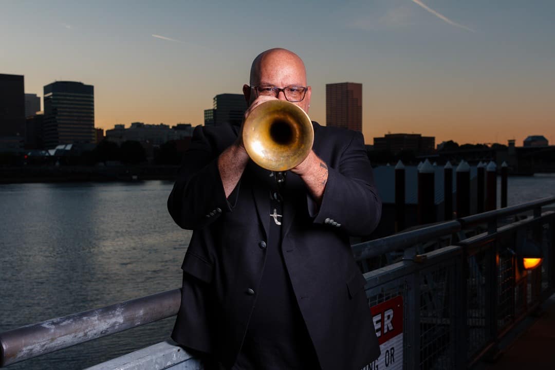 Bald man playing trumpet outdoors at sunset in an urban waterfront setting for music, jazz, or performance themed image.
