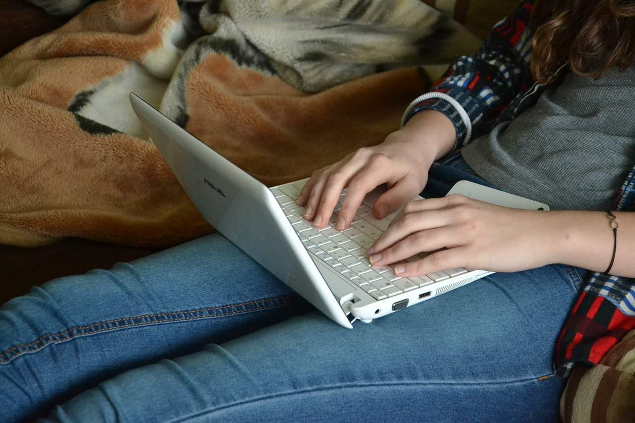 Laptop computer on person's lap, casual indoor setting, young woman typing, cozy environment, tech stay-at-home working or learning, soft blanket background, modern digital device, home office setup, casual lifestyle image, everyday tech use relevance.