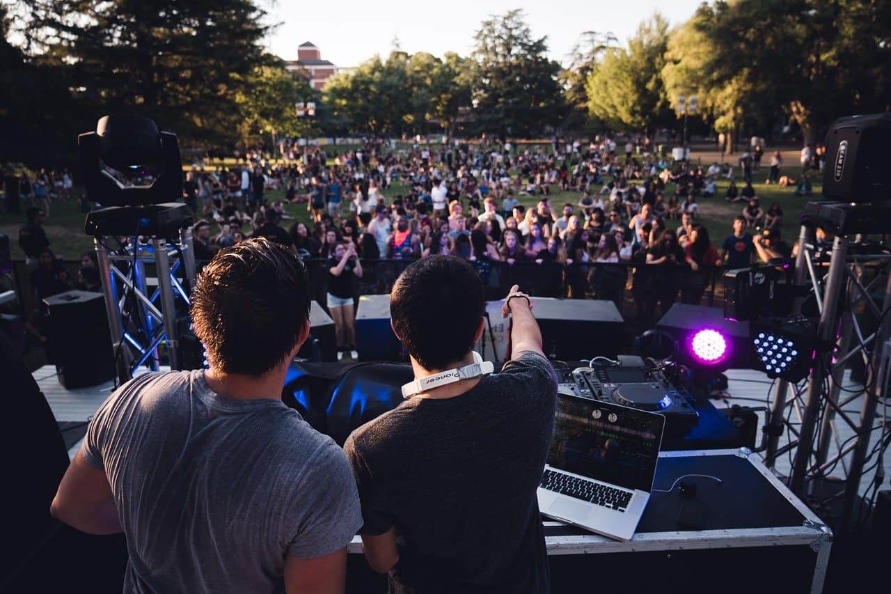 VJ DJs performing at an outdoor music festival with a large crowd, stage lighting, and DJ equipment in the foreground, capturing the vibrant electronic dance music scene and live event atmosphere.