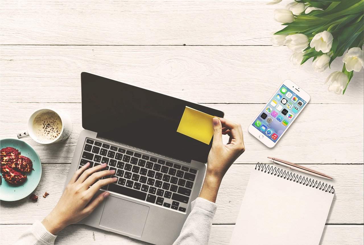 Laptop with sticky note and smartphone on wooden desk, workspace for digital content creation, social media, and music industry research, with flowers and snacks.