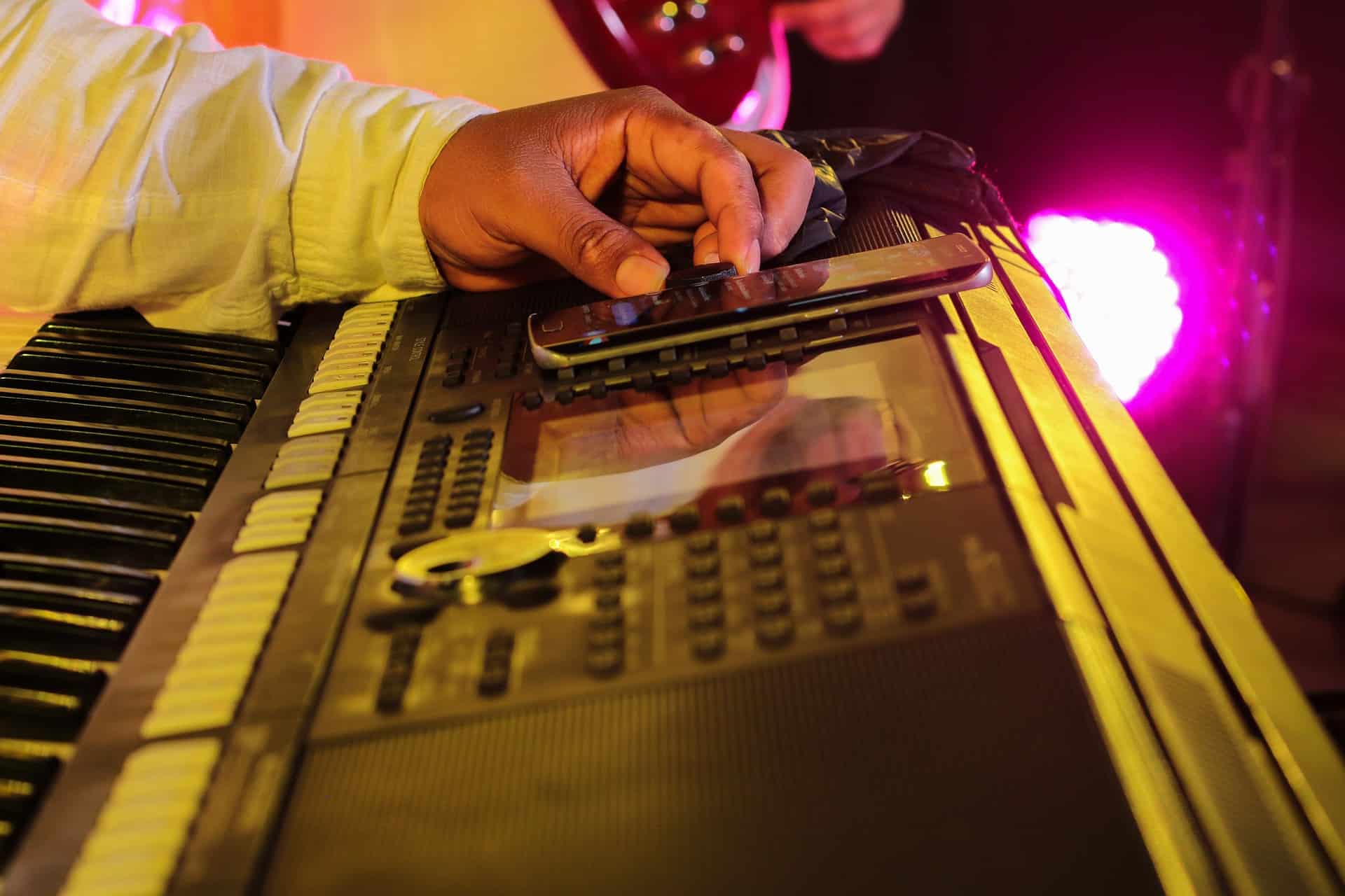 Vocalist playing a keyboard with a smartphone on top during a musical performance under colorful stage lighting.