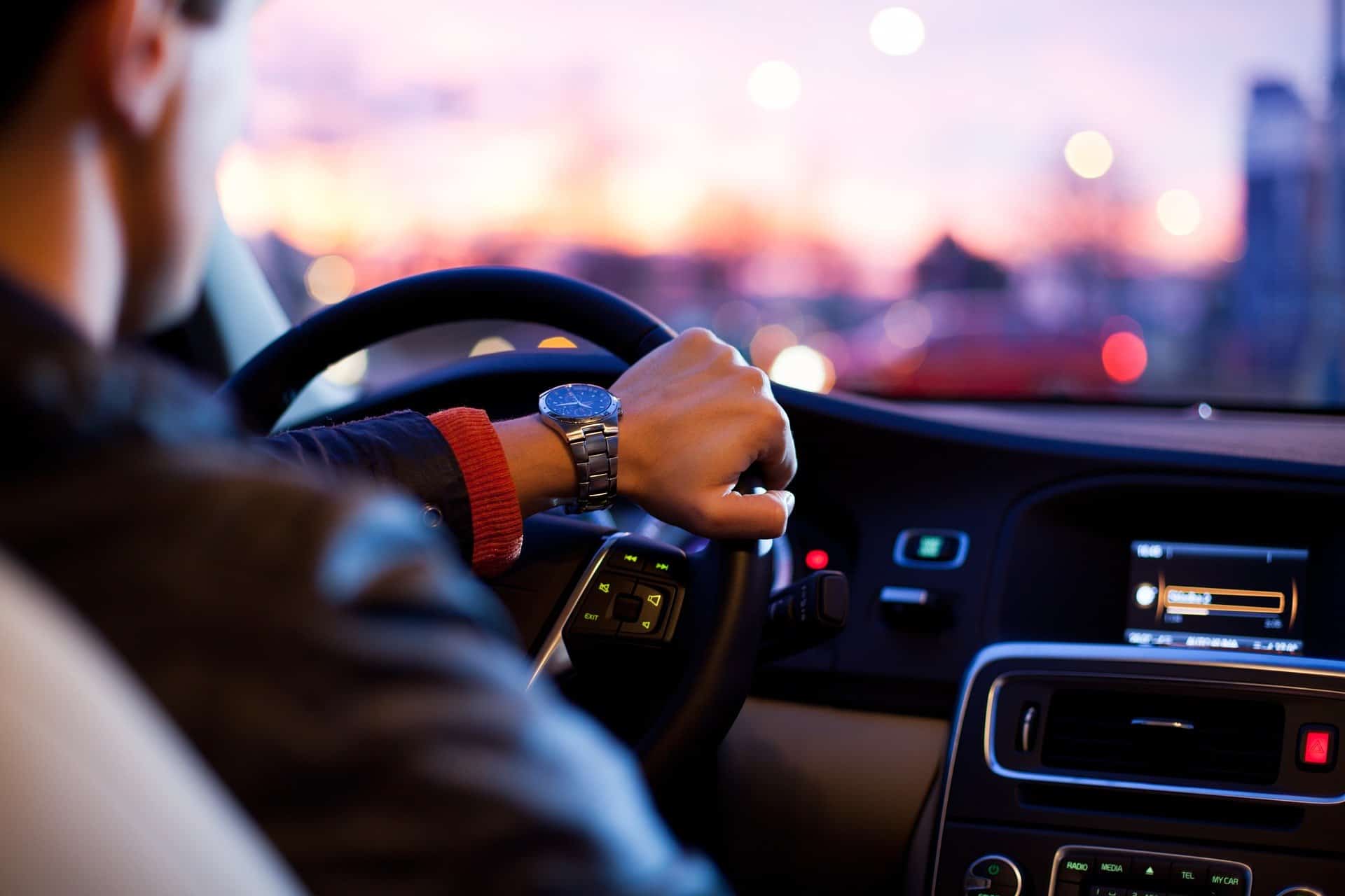 A person driving a car at sunset, showing a close-up of their hand on the steering wheel and wearing a wristwatch, with a blurred cityscape outside.