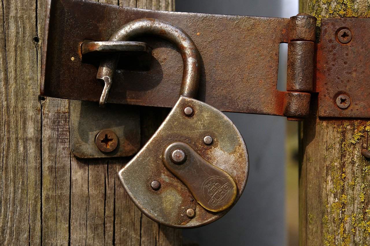 Locked padlock securing wooden gate, rusty metal latch and screws, outdoor security, weathered wood, padlock lock, outdoor safety, mechanical security, metal hardware, rustic security detail, outdoor gate lock, metal latch and key, traditional locking device, outdoor environment, security hardware, weathered outdoor lock.