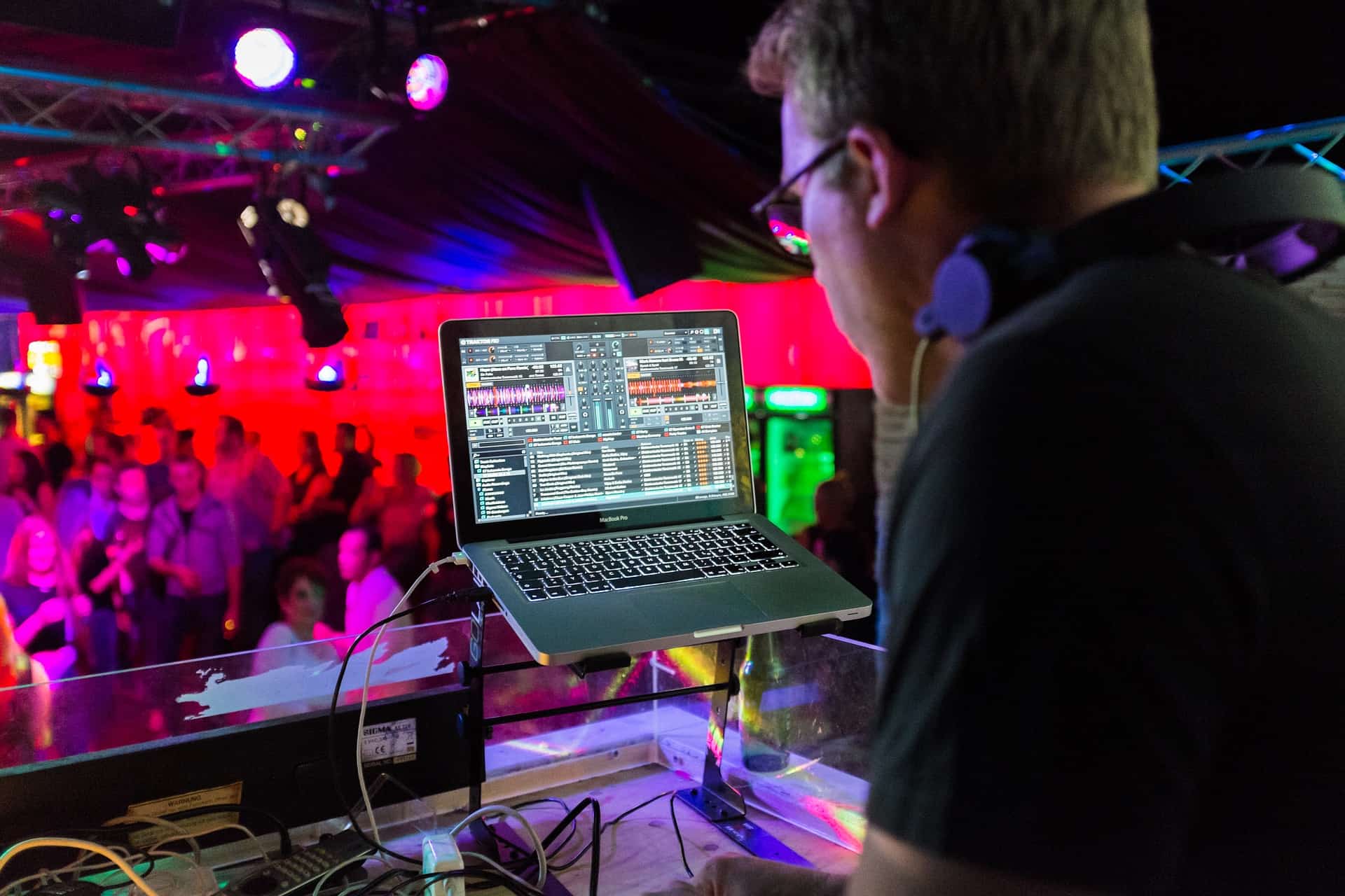 DJ mixing music on a laptop at a lively nightclub or concert venue with colorful stage lighting and dancing crowd.