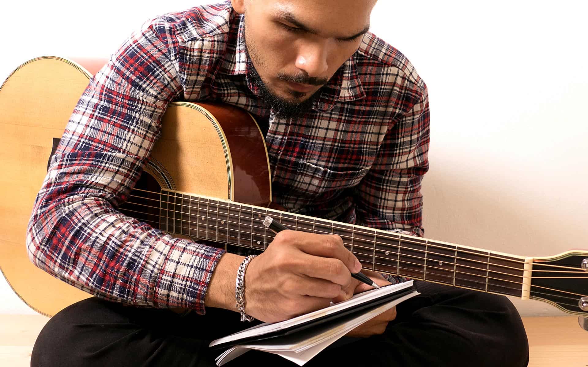 Guitar player writing notes with a pen on a notepad, sitting indoors. Focus on musician engaged in songwriting or practice with acoustic guitar, highlighting music creativity, composition, and artistry.