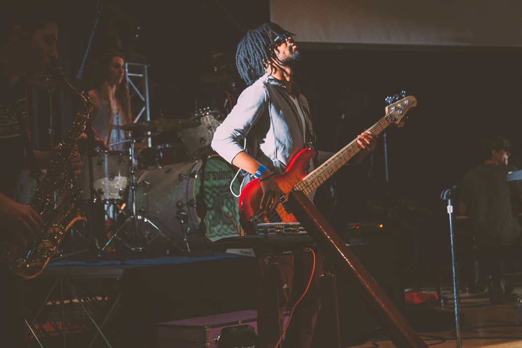 Vibrant live music performance featuring a bass guitar player with dreadlocks, a drummer, and saxophonist on stage, capturing the energy of a concert or music event.