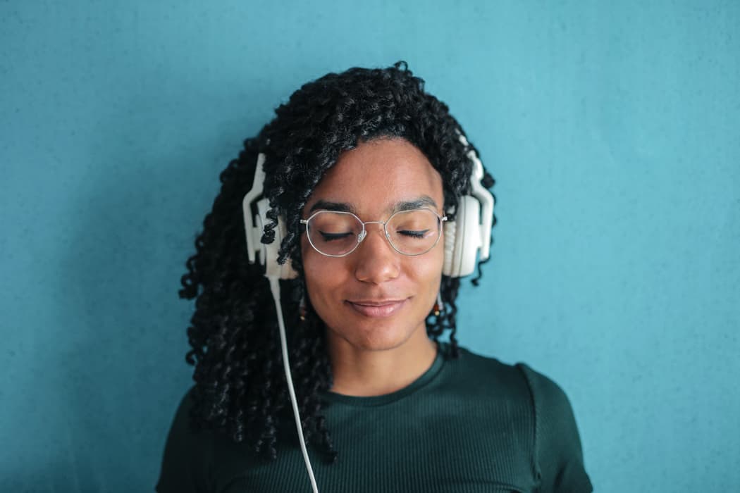 Relaxed woman listening to music with headphones, wearing glasses and a dark shirt, standing against a blue background.