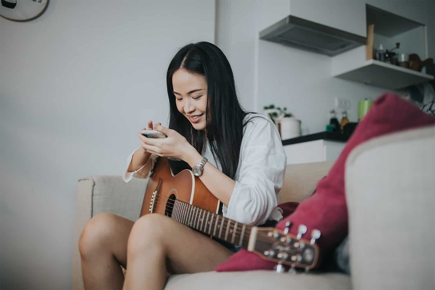 Young woman playing acoustic guitar while using smartphone at home, creative music practice, casual lifestyle, relaxed atmosphere.