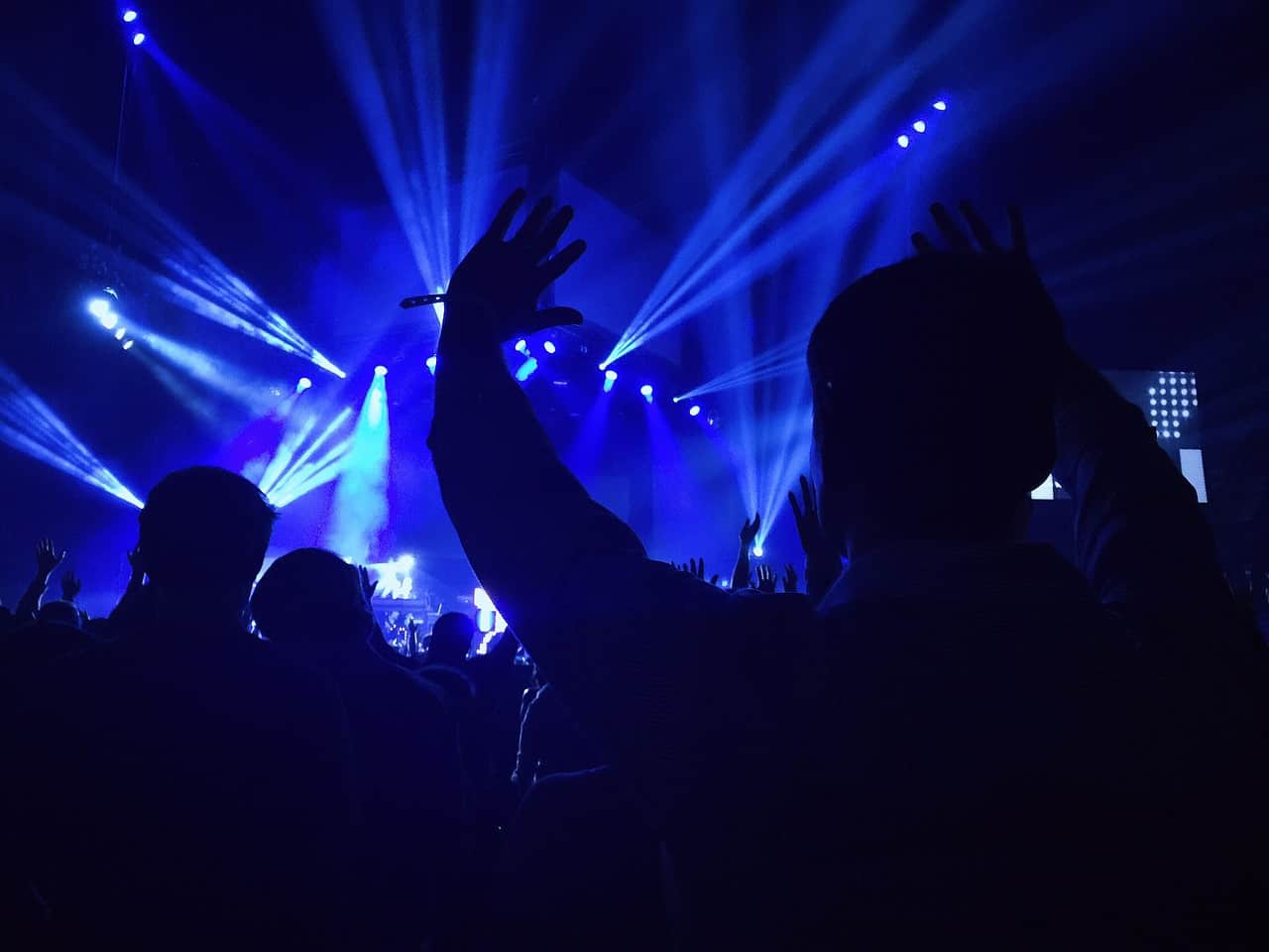 Vibrant concert scene with silhouettes of audience members dancing and raising hands under dynamic blue stage lighting, highlighting live music, entertainment, and event atmosphere.