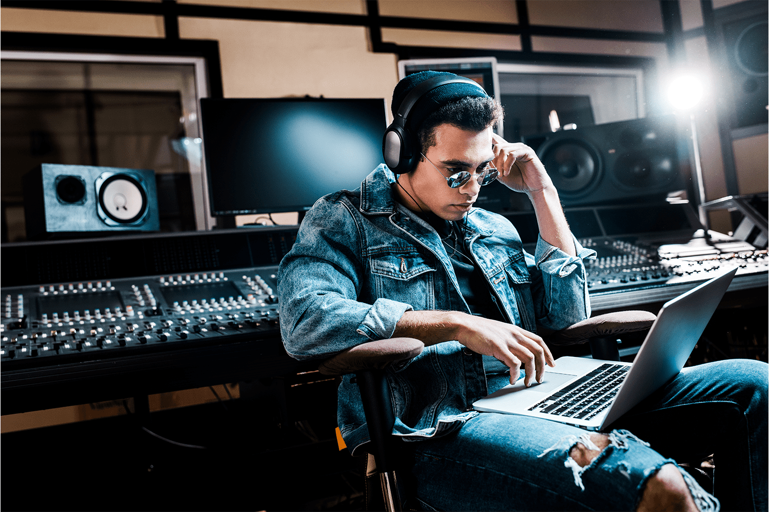 Young male music producer working in a professional recording studio with audio mixing equipment, headphones, and a laptop, focused on creating and mixing music tracks.