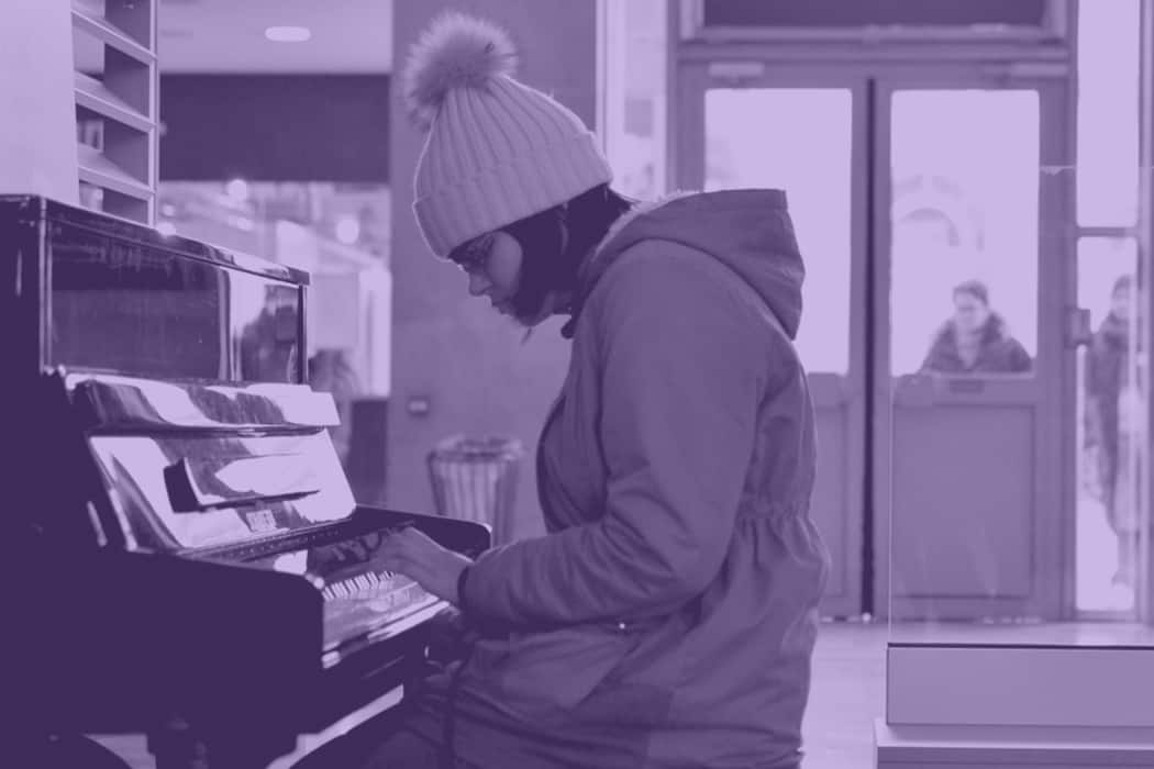 Young woman wearing winter hat and glasses playing piano in a public space, music or busking performance, urban setting.