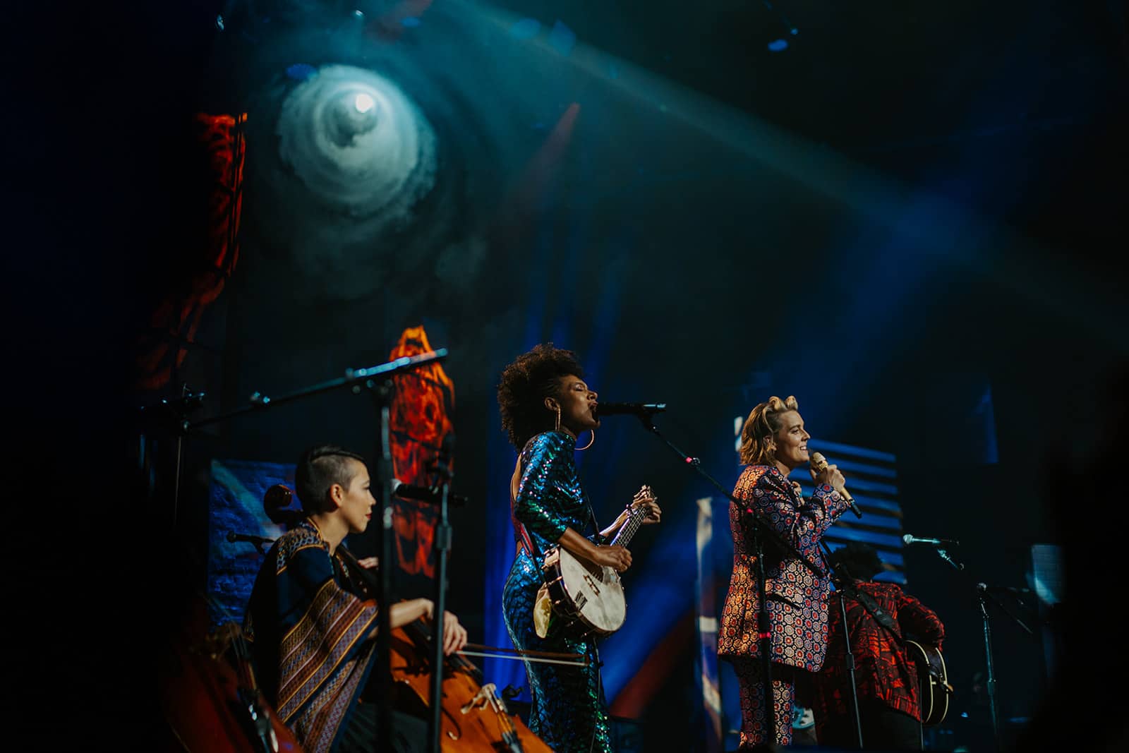 Vibrant live musical performance featuring female singers and musicians on stage with dramatic lighting and colorful backdrop, capturing the energy of a concert or music event.