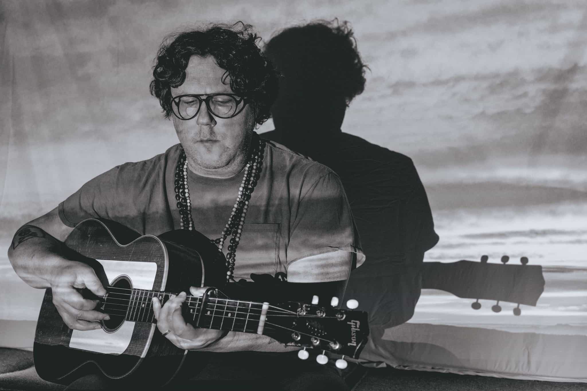 Soft-focus black and white image of a man with curly hair and glasses playing an acoustic guitar, with a sunset backdrop, capturing a mood of musical inspiration and artistic expression.