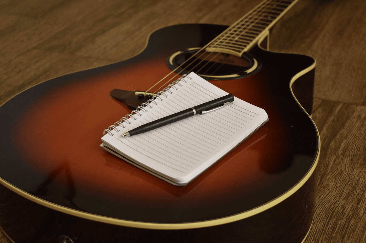 A photo of an acoustic guitar with a notepad and pen on top, placed on a wooden floor, symbolizing music creation, songwriting, and musical inspiration.