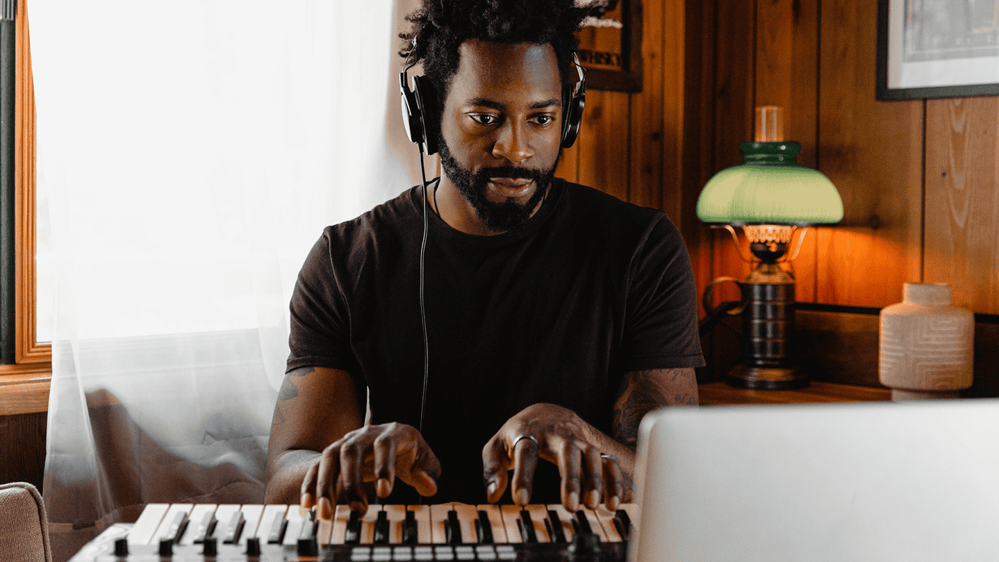 Music producer working on a MIDI keyboard with headphones, creating music in a cozy, wooden-paneled home studio environment.