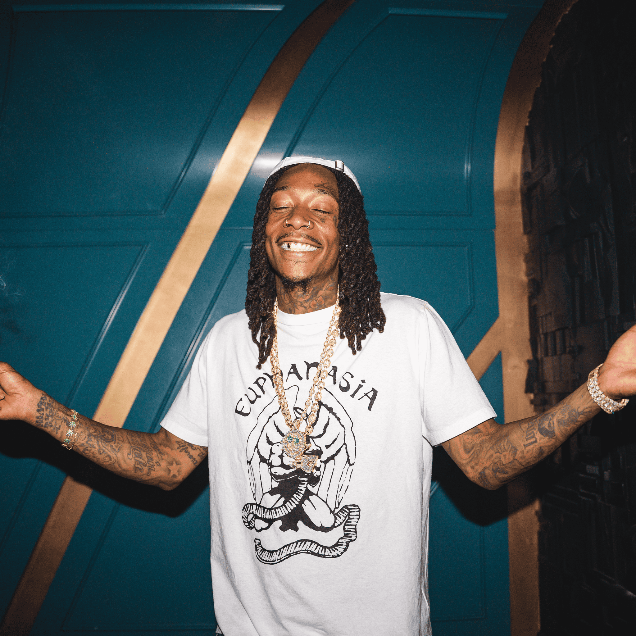 Hip-hop artist with long dreadlocks wearing a white graphic T-shirt and gold jewelry, smiling with arms wide open against a modern blue and gold wall background, music and entertainment, urban culture, artist portrait.