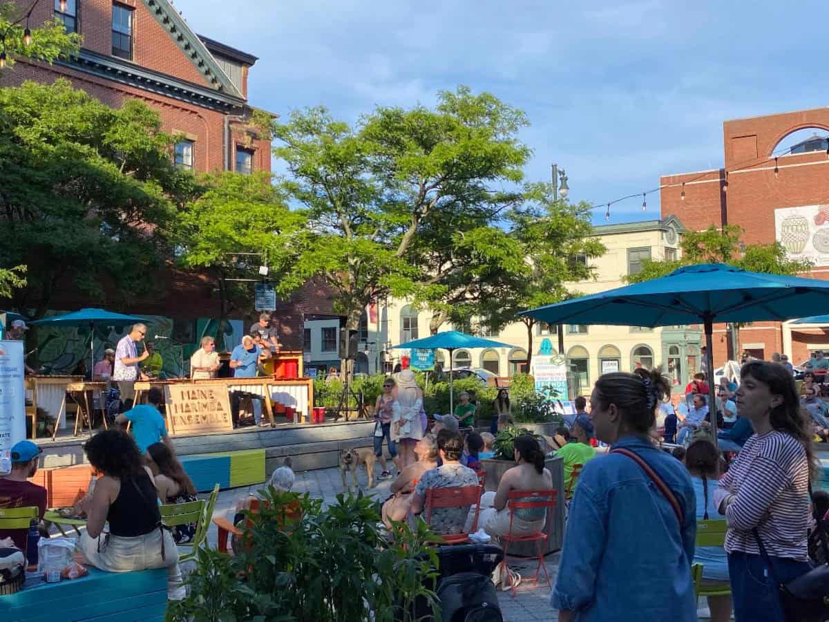 Vibrant outdoor summer concert in an urban park with diverse audience, musicians performing on stage, lush trees, colorful seating, and city buildings in the background, capturing community engagement and live music.