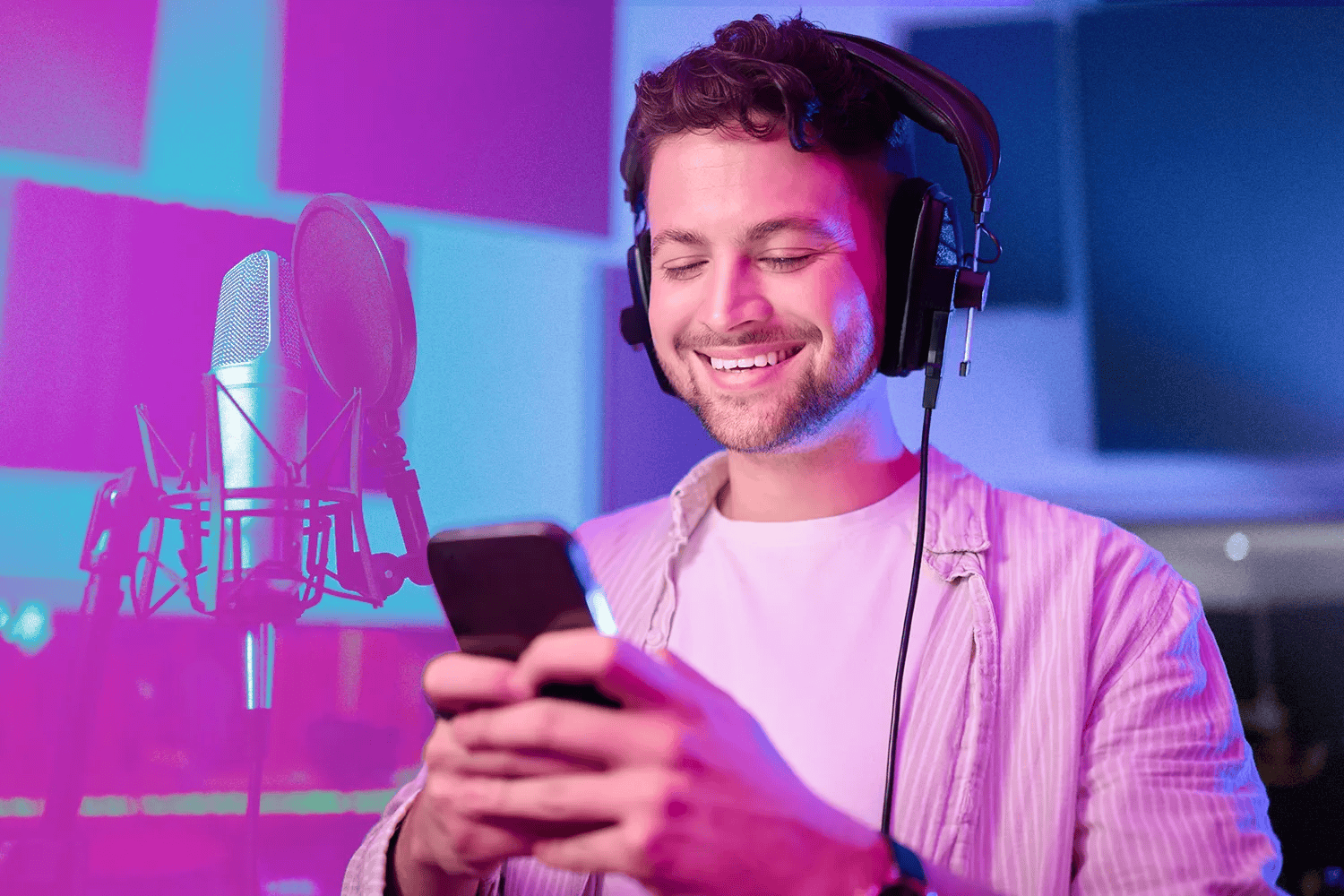 Young man in a recording studio using headphones and smartphone, with microphone setup, focusing on music production, digital content creation, and audio recording.