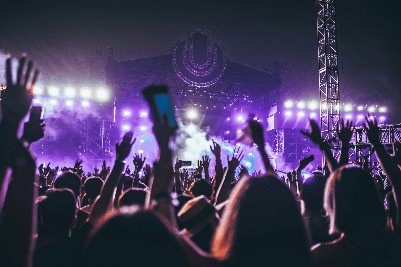 Vibrant music festival with crowd dancing under purple stage lights, large U-shaped logo on stage, energetic concert atmosphere, live performance, electronic dance music scene, night event.