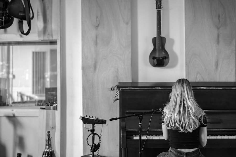 A person with long hair playing piano in a music studio with a guitar hanging on the wall.