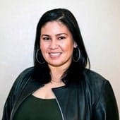 A confident woman with dark hair wearing a black leather jacket, smiling against a light background.