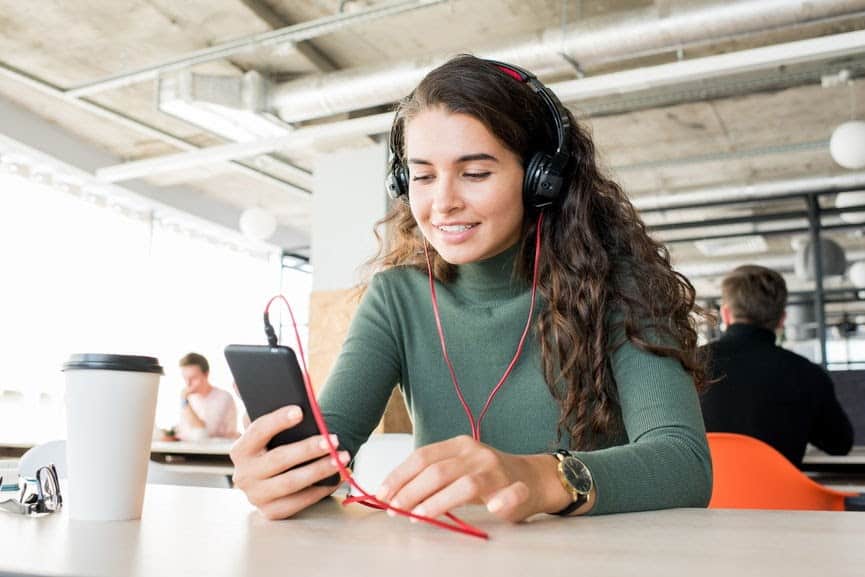 Young woman with headphones using smartphone in a modern coffee shop, enjoying music or a podcast, with a coffee cup on the table, casual workspace, vibrant and relaxed atmosphere.