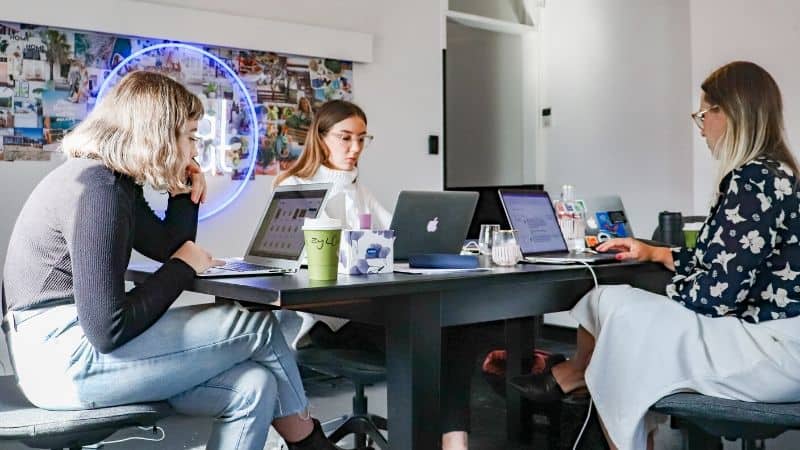 Laptops and notebooks on a conference table during a team meeting or brainstorming session with three women working together in modern office space.