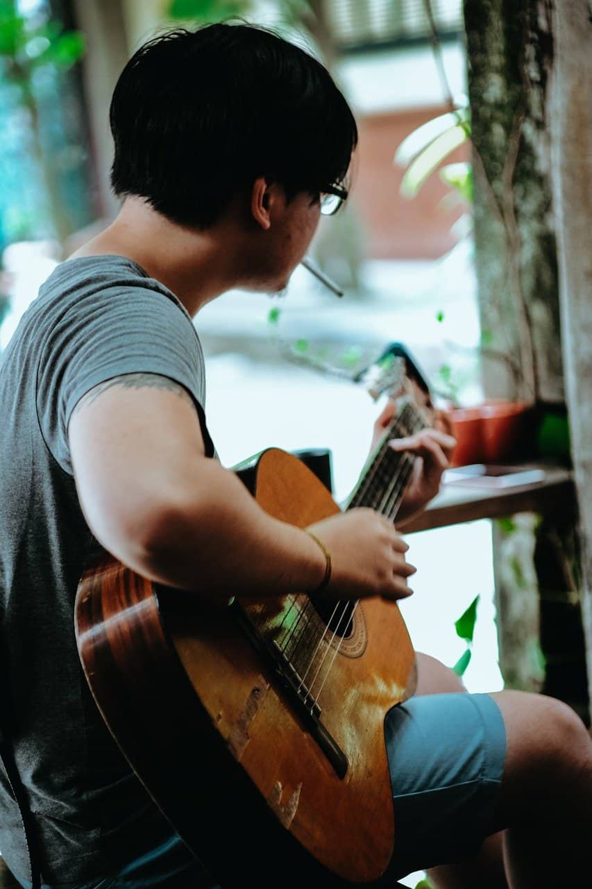 Guitar player playing acoustic guitar, casual musician practicing music, outdoor setting with greenery, young man focused on guitar, music and arts, hobby and leisure activities, street music scene, creative expression, personal music journey, artistic lifestyle.