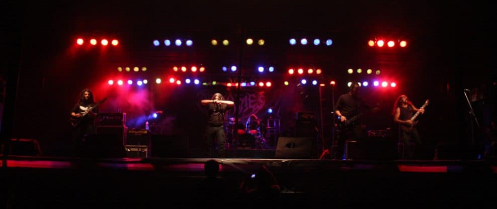 Guitarists and drummer performing live on stage with vibrant red, blue, and yellow stage lighting, showcasing a dynamic concert atmosphere.