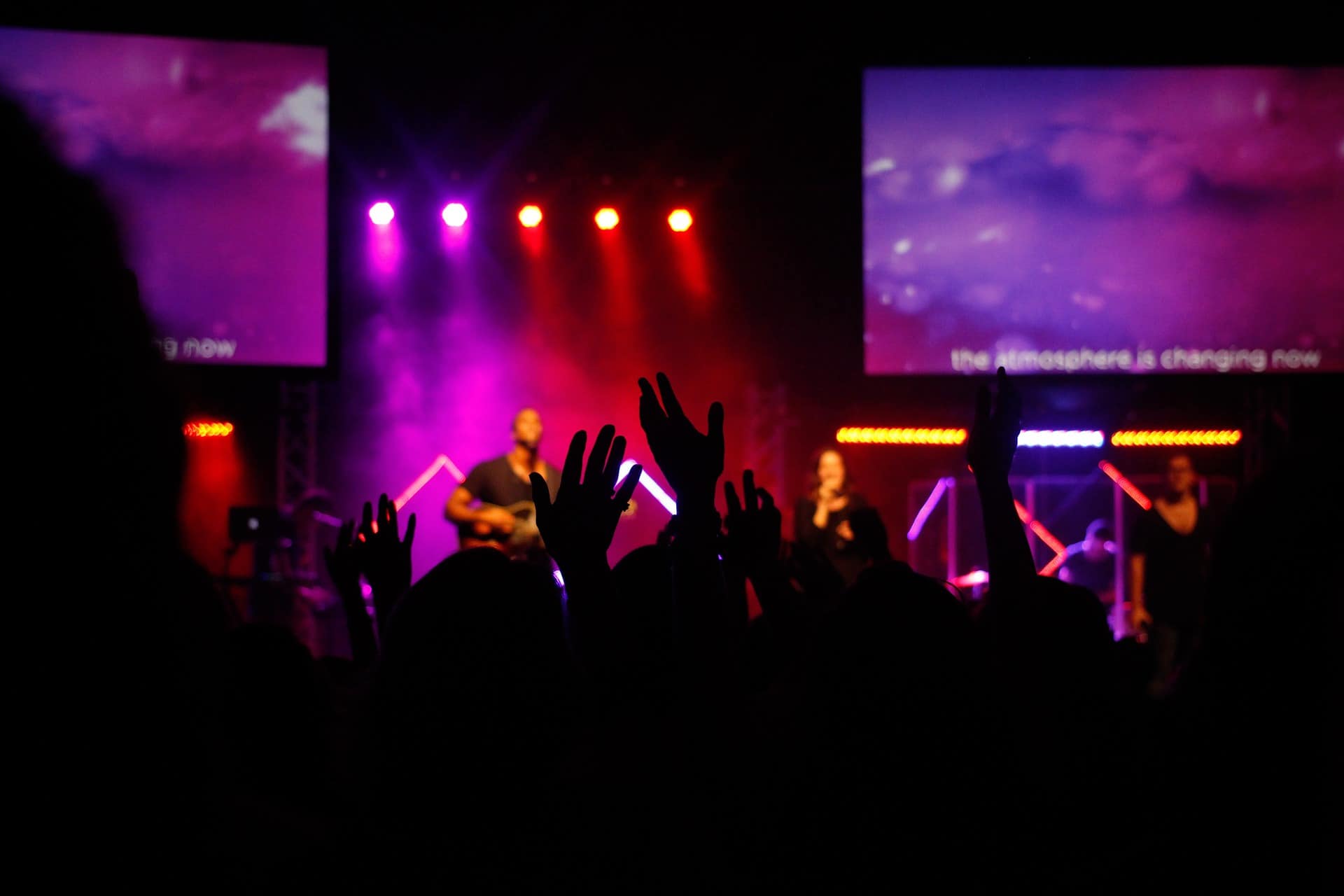 Concert with live music, audience raising hands, vibrant stage lighting, worship band performing, colorful purple and red lights, church or music event atmosphere, energetic crowd scene, multimedia screens in background, modern concert environment, music performance visuals, engaging worship experience, event photography, nightlife entertainment, worship music concert, stage illuminated with purple and pink lighting.