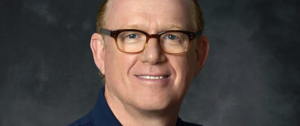 A professional headshot of a smiling man wearing glasses and a dark blue shirt, suitable for music industry or digital media content.
