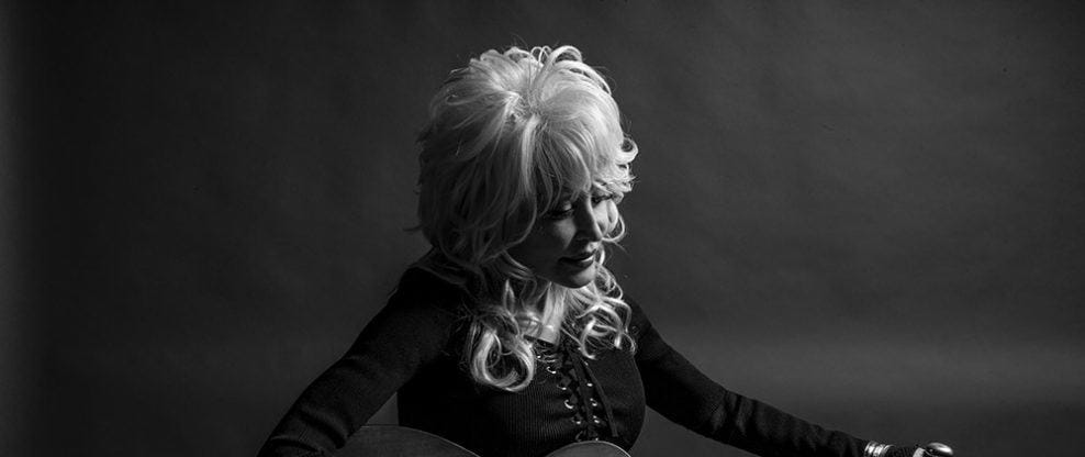 Black and white portrait of a woman with blonde hair in a classic style, dressed in a black top, looking thoughtfully downward against a dark background.