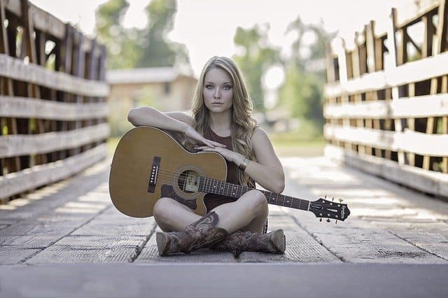 Acoustic guitar player woman sitting outdoors on wooden bridge with guitar, casual music artist, young female musician, singer-songwriter, outdoor performance, youth culture.