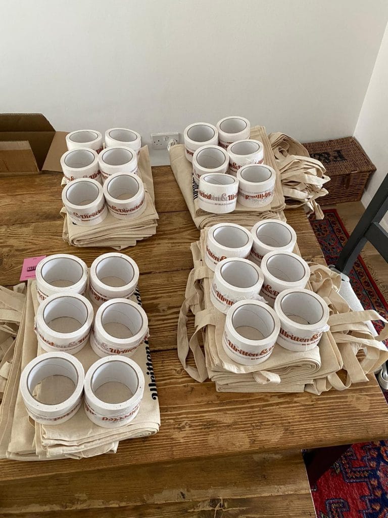 Wristbands and tote bags for a promotional event or concert, organized on a wooden table, with a plain white wall background, emphasizing brand merchandise distribution.