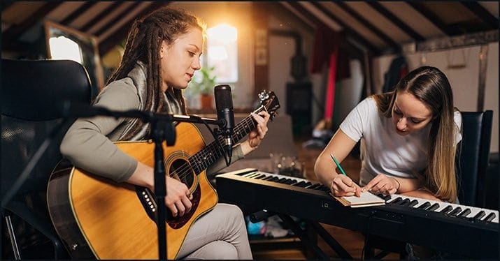 Young women musicians practicing in a cozy attic studio, one playing guitar and the other composing on a keyboard, embodying creativity and musical talent.