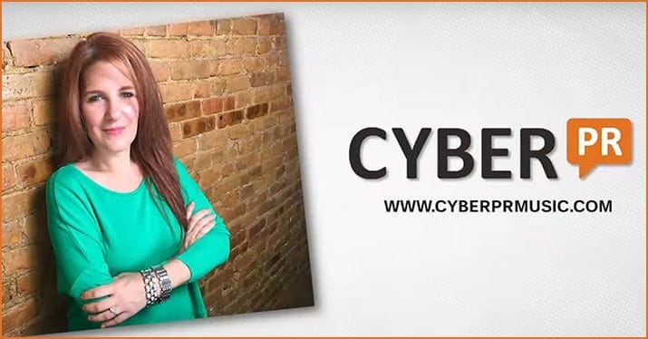 Girl with red hair in a green sweater standing against a brick wall for Cyber PR music promotion at Hypebot.
