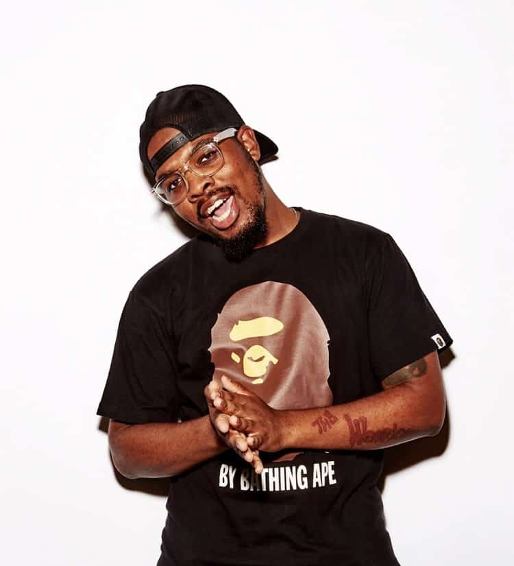 Fashion influencer wearing Bape graphic t-shirt, stylish baseball cap, and glasses, posing against white background for streetwear and hip-hop fashion content.