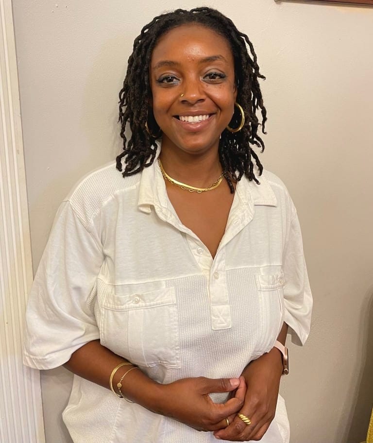 Beautiful woman with dreadlocks smiling, wearing a white casual shirt, gold jewelry, and standing indoors, representing confidence and style.