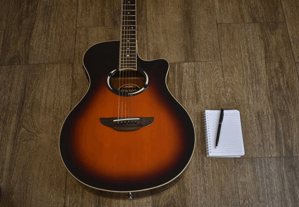 Guitar with notepad and pen on wooden floor, music practice, songwriting, acoustic guitar, creative process, music industry, musician tools, musical inspiration, Hypebot feature.