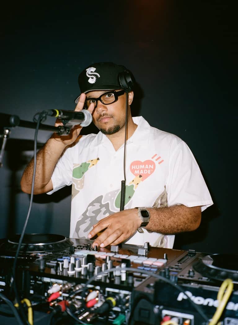DJ performing live with turntables and mixer, wearing headphones and a branded cap, promoting music production and DJ culture at a music event.