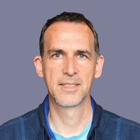 Professional portrait of a man with short dark hair and blue eyes, wearing a blue jacket against a plain grey background.