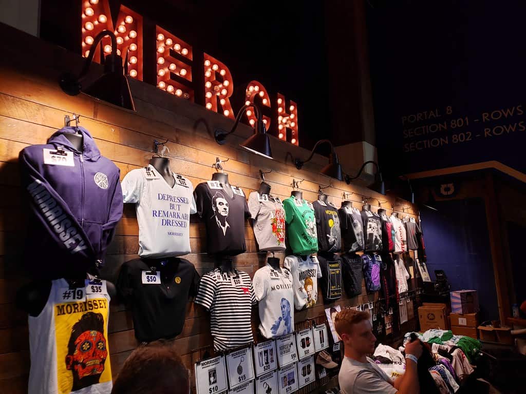Vintage rock band merchandise display in apparel store with Morrissey T-shirts and accessories, illuminated by retro signage and warm lighting, showcasing music memorabilia and fan merchandise.